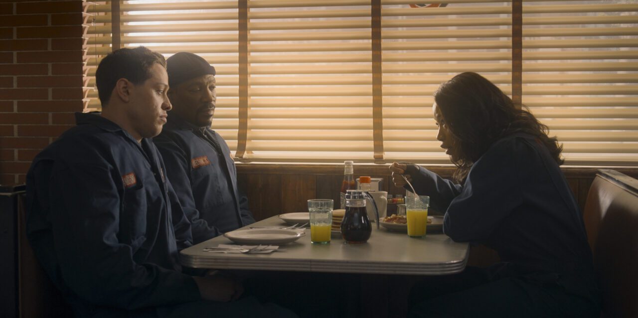 Three individuals, two men in what appear to be work uniforms and a woman eating, seated in a diner booth with venetian blinds on the windows, from the movie The Pickup.
